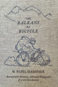 Book cover of The Balkans By Bicycle by W Papel Hamsher featuring a cartoon of the author astride his bicycle.