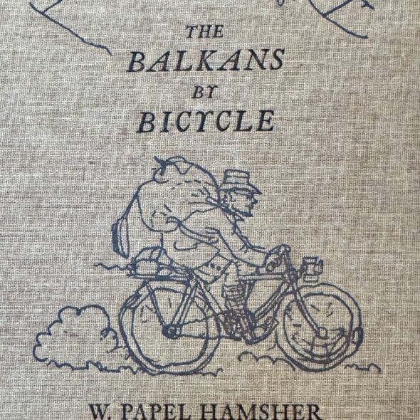 Book cover of The Balkans By Bicycle by W Papel Hamsher featuring a cartoon of the author astride his bicycle.